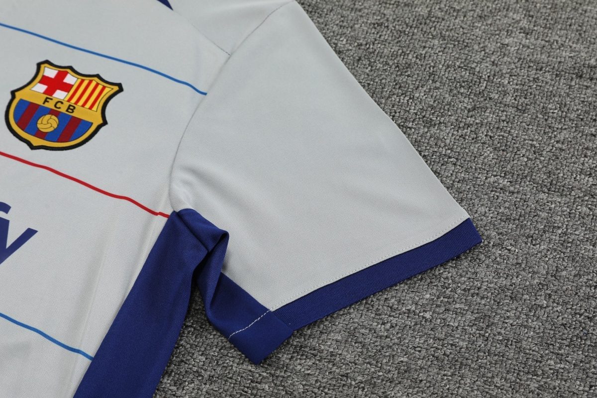 Partially folded white FC Barcelona football jersey on a gray carpet, showing the club crest and blue trim.