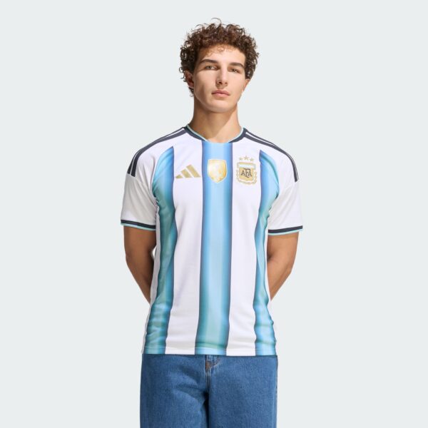Young man wearing an Argentina national team jersey, standing against a pale gray background.