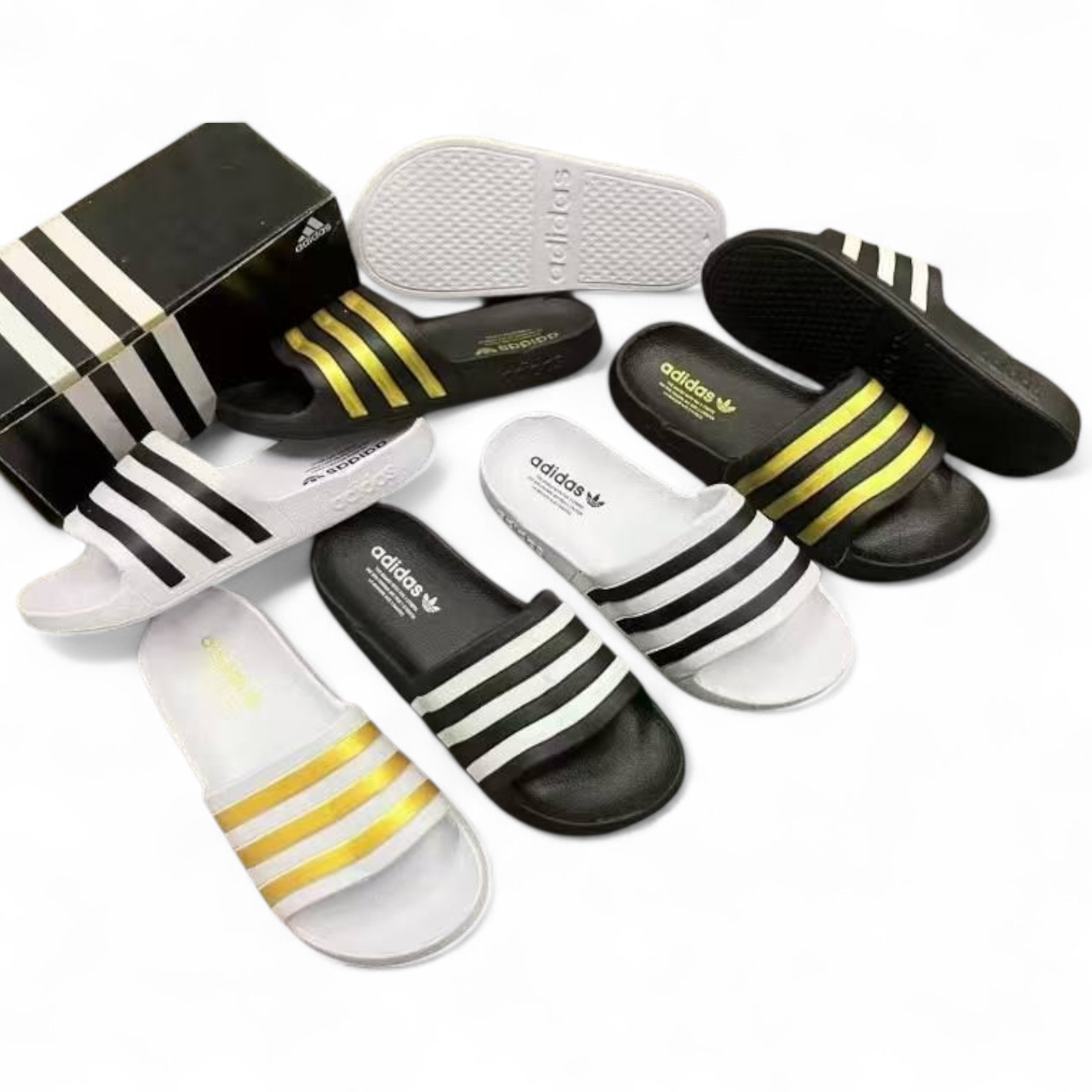 rn-image_picker_lib_temp_e0a9af7a-53bb-4dce-9f95-e3dc143591d5 Assorted Adidas slide sandals in black and white with yellow stripes, arranged around a black Adidas box.