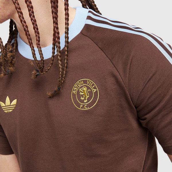 Close-up of a person wearing a brown Aston Villa FC jersey with a light blue collar and stripes, and braided hair visible beside the chest crest.