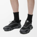 Person wearing black and gray chunky sneakers with black socks, standing on a light background (product photo).