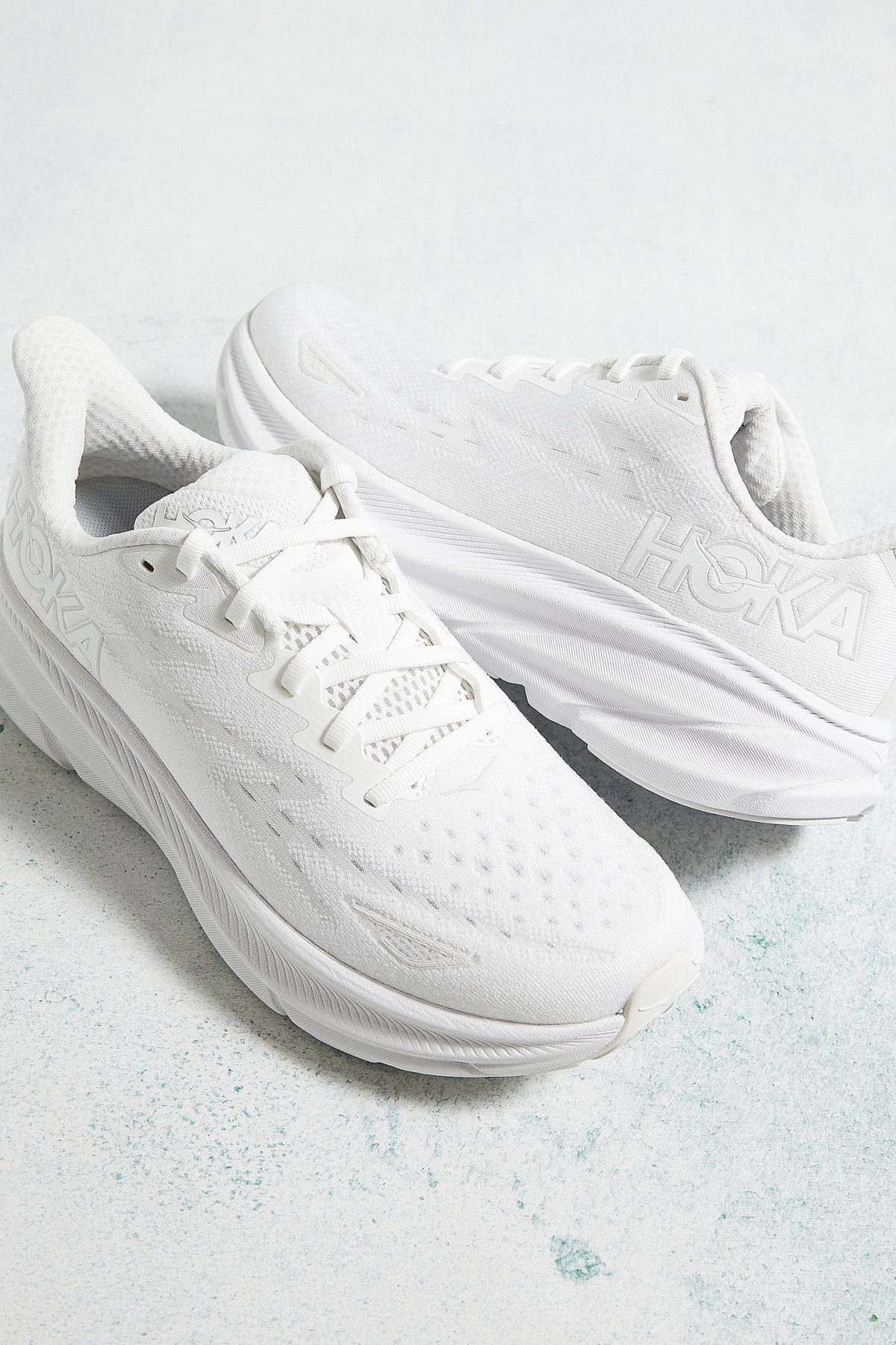 Pair of white athletic sneakers with laces on a pale, textured surface.