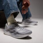 rn-image_picker_lib_temp_eac3c08a-8af0-49ca-adb8-e89817b0e8ee | UAE SPORT STORE Gray athletic sneakers on a person wearing denim jeans and beige socks, feet resting on the floor in a studio setting