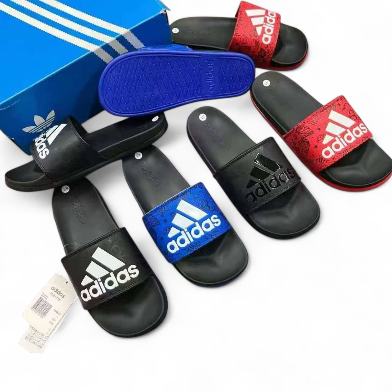rn-image_picker_lib_temp_ec230462-4b9b-4b34-ac96-1fcf2cc9e01d Assorted Adidas slide sandals in black, blue, and red with white logos, arranged near a blue shoe box.