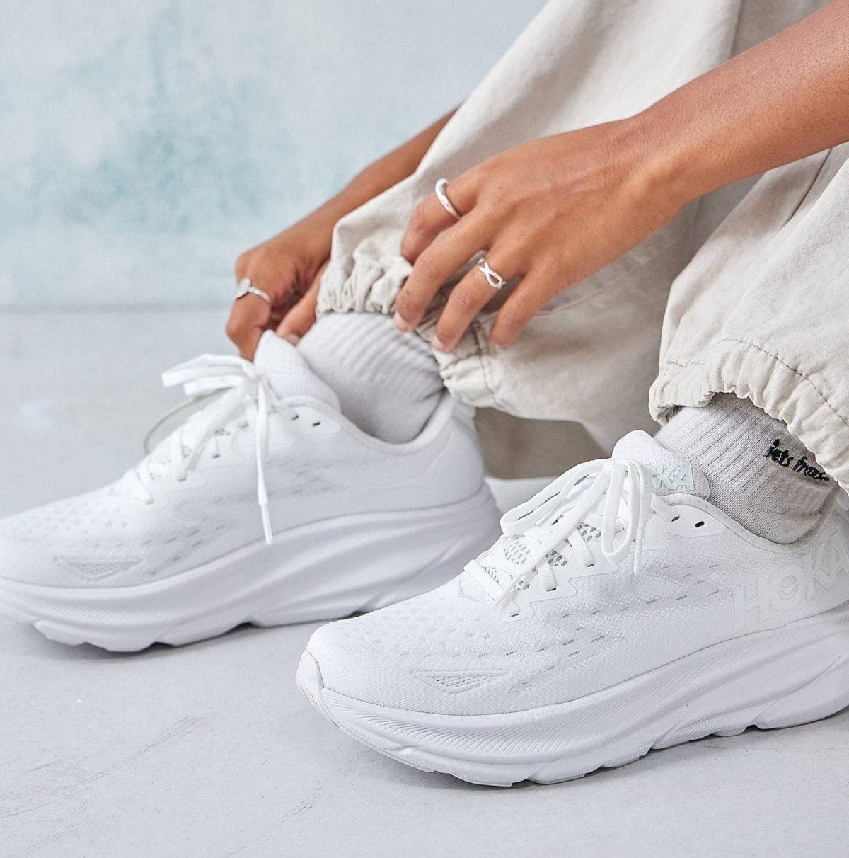 Close-up of a person tying the laces on white athletic sneakers, with silver rings on their fingers and light gray joggers nearby.