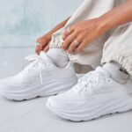 Close-up of a person tying the laces on white athletic sneakers, with silver rings on their fingers and light gray joggers nearby.