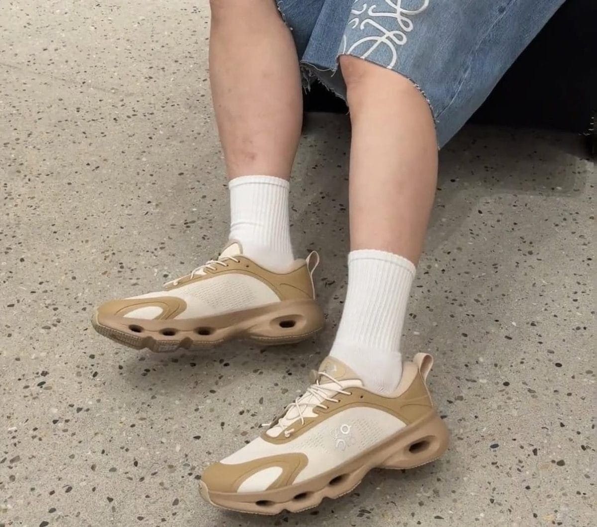 Beige and cream athletic sneakers with white socks on a speckled floor.
