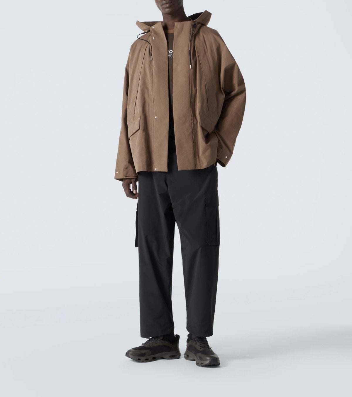 Full-body view of a person wearing a brown hooded jacket, black cargo pants, and brown sneakers, standing against a light background.
