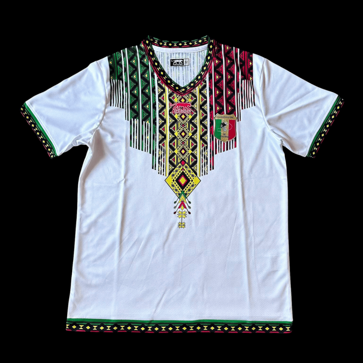 White short-sleeve dashiki with a multicolored geometric pattern around the neckline and fringe tassels; green trim on sleeves and hem.