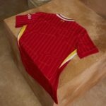 Red sports jersey with thin yellow geometric lines, white and yellow side panels, laid flat on a brown stone block (viewed from above). Note the Adidas branding on the sleeve edge.