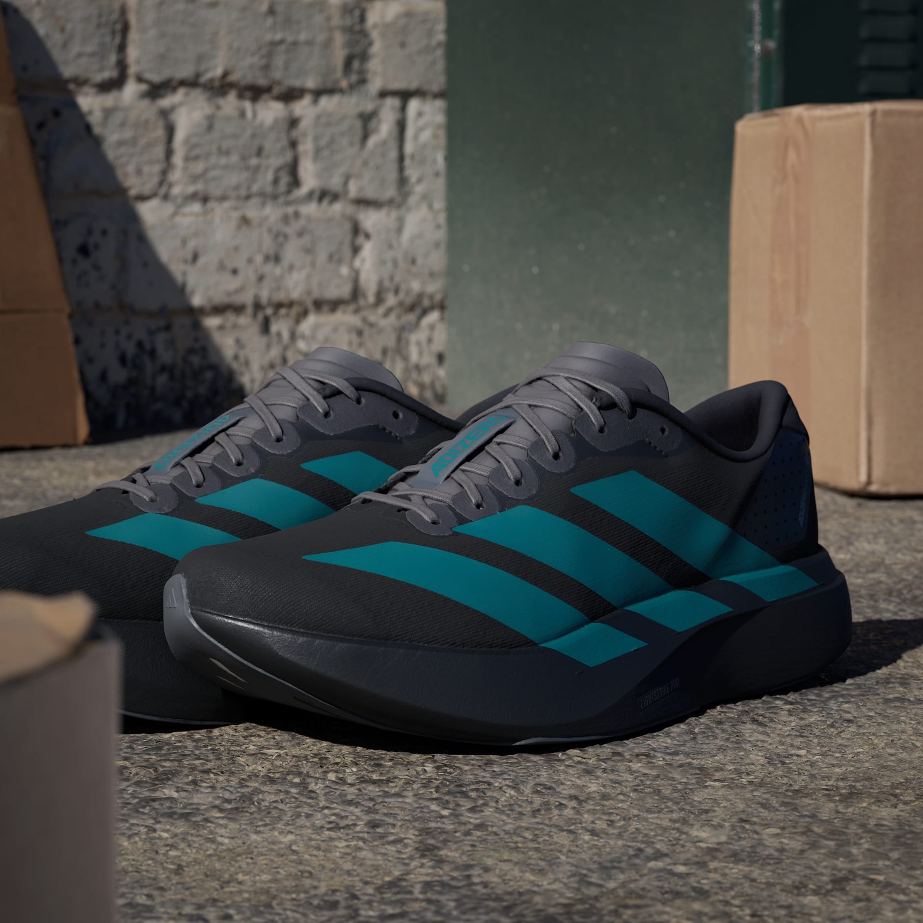 rn-image_picker_lib_temp_f7bd889c-6564-4413-a7ed-f1c32c1e618d Close-up of gray running shoes with teal stripes resting on rough outdoor ground with a brick wall and cardboard boxes in the background