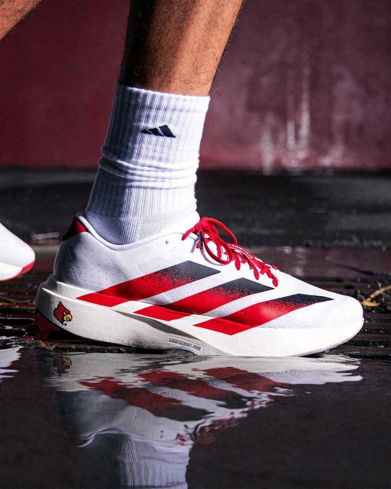 rn-image_picker_lib_temp_fdeb8ae5-9097-4216-b612-40dbdaa5646b | UAE SPORT STORE White athletic shoe with red stripes and a Louisville Cardinals logo on the heel, standing on a wet, reflective surface.