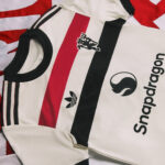 White athletic jersey with red and black vertical stripes, Adidas logo, and sponsor text along the side.