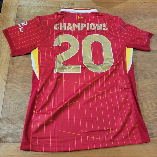 Red soccer jersey laid flat, back showing 'CHAMPIONS' and number 20 in gold, with yellow trim and geometric orange lines.