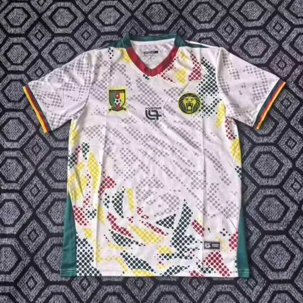 White Cameroon national soccer jersey with multicolor dotted pattern, green side panels, and striped sleeve cuffs; crest on the left chest and a yellow lion emblem on the right.