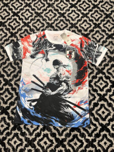 Graphic t-shirt featuring a stoic samurai with two swords in a bold black, white, red, and blue illustration, and a Japan JFA patch on the chest, laid on a black-and-white patterned rug.