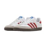 White Adidas sneakers with blue tongue logo, red stripes, and beige suede toe cap on a brown sole