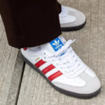 White Adidas Samba sneaker with red stripes on a wooden surface, laces tied, pant leg covering the ankle.