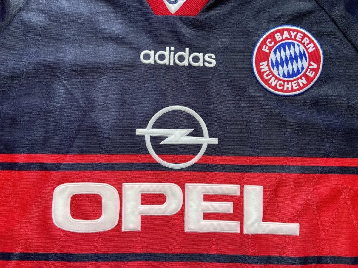 Dark blue FC Bayern jersey with Adidas logo, Opel emblem, and club badge visible on chest; red sponsor band at bottom reading OPEN partially.