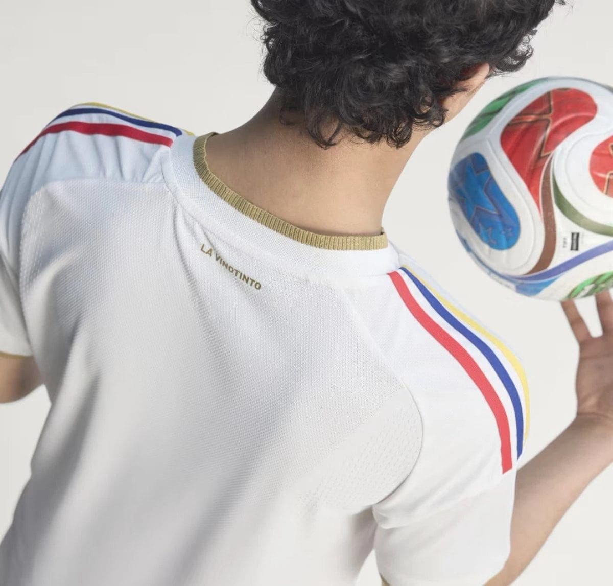 Back view of a person wearing a white soccer jersey with gold collar and red, blue, and yellow shoulder stripes, holding a colorful soccer ball to the right.