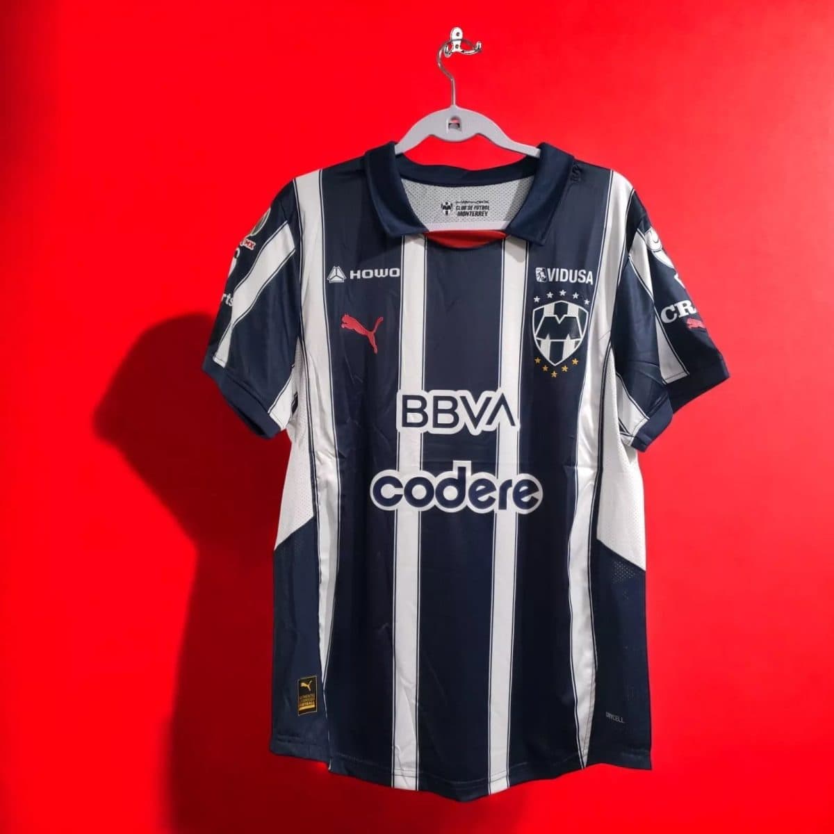 Navy and white striped football jersey hanging on a hanger against a red background, with crest and BBVA sponsor.