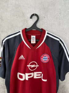 FC Bayern Munich soccer jersey on a black hanger against a concrete wall; red center with navy sides and Opel sponsor logo visible.