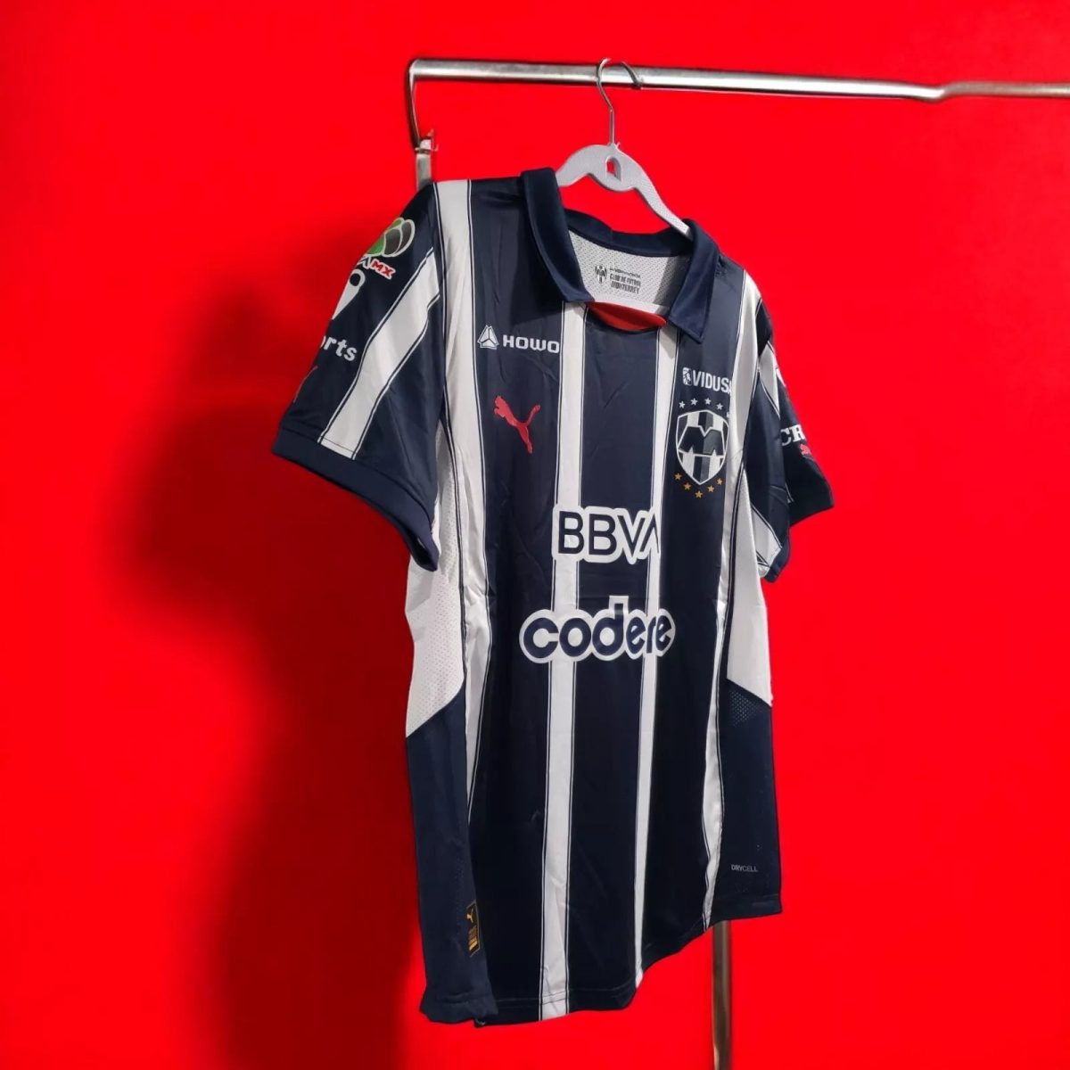 Blue and white vertical striped soccer jersey hanging on a hanger against a bright red background, with sponsor logos visible on the chest area (BBVA).