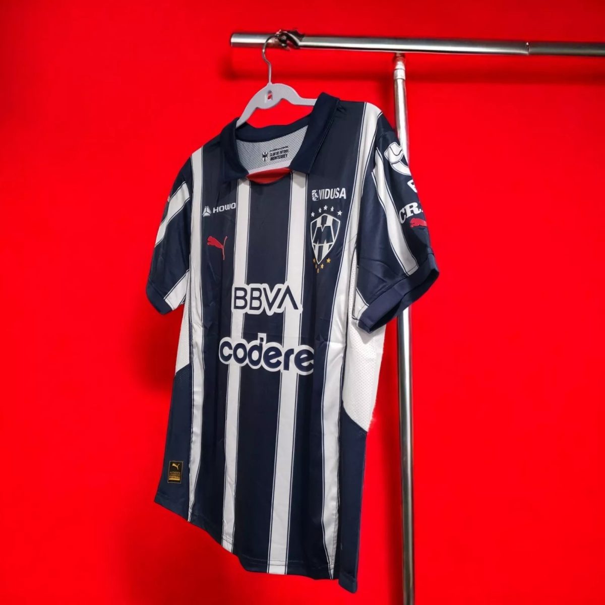Navy and white striped soccer jersey on a hanger against a bright red wall, showing sponsor logos.
