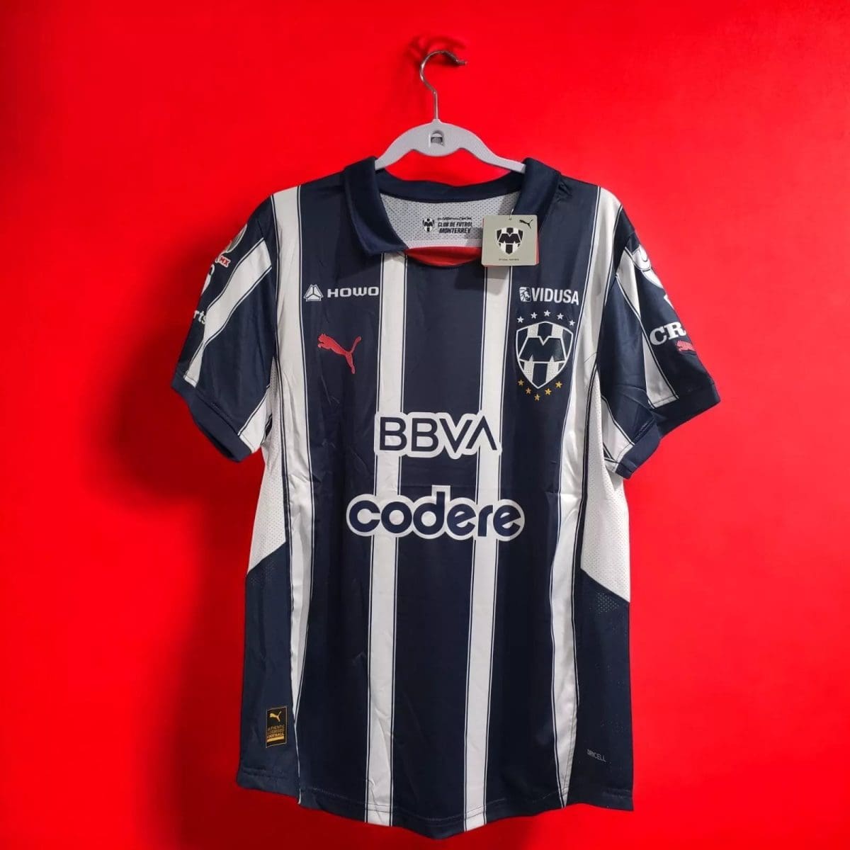 Navy and white striped soccer jersey on a hanger against a bright red wall, with club crest and sponsor logos (BBVA, Codere).