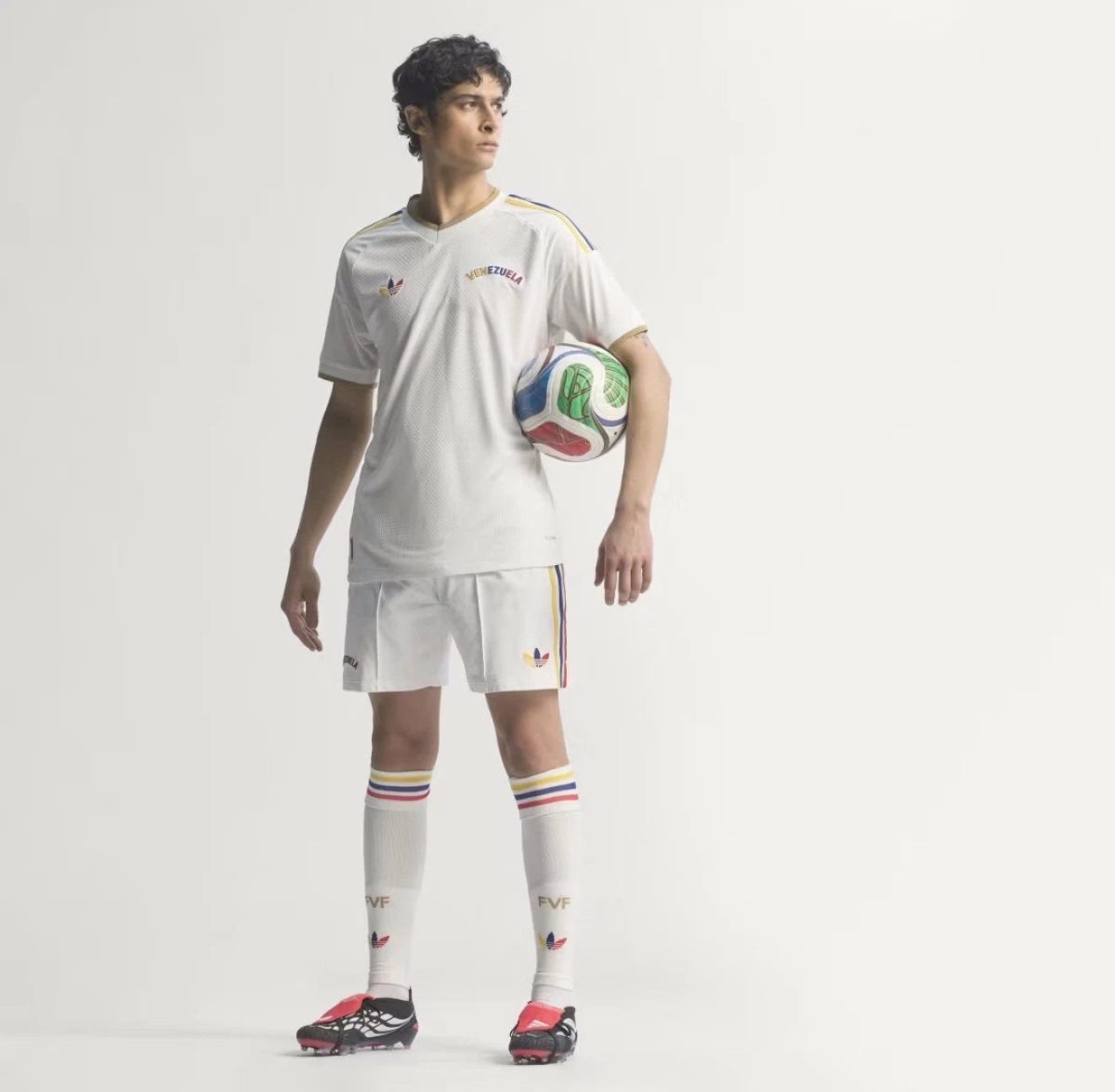 Male soccer player in a white Venezuela kit standing with a multicolored soccer ball under his arm against a light background.
