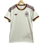 White Spain national football jersey with maroon Adidas stripes on shoulders and gold trim, crest on left chest, hanging on a wooden hanger.