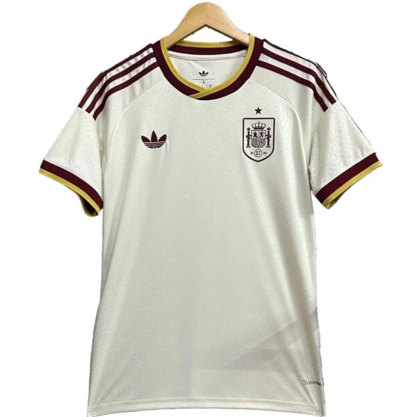 White Spain national football jersey with maroon Adidas stripes on shoulders and gold trim, crest on left chest, hanging on a wooden hanger.
