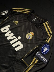 Black Real Madrid jersey with gold stripes and sponsor logo, featuring the crest, Adidas emblem, and patch details (UEFA badge and Respect sleeve patch).