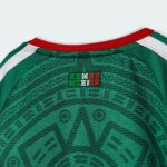 Close-up of a green jersey back with a red collar and white shoulder stripes, featuring a small Somos Mexico logo in the center.