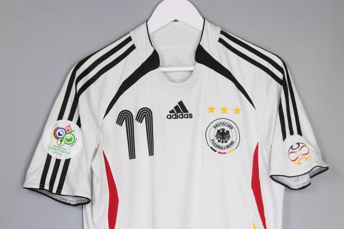 White Germany national team football jersey on a hanger with number 11 and three gold stars above the crest, Adidas logo, and World Cup 2006 badge on the sleeve edge.