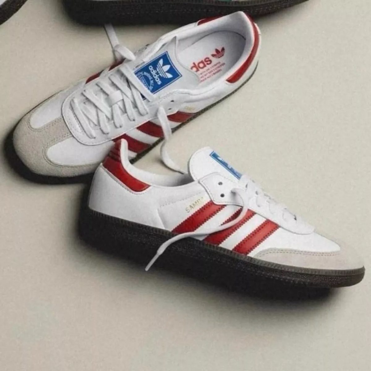 Pair of white sneakers with red Adidas stripes and black rubber sole resting on a light surface.