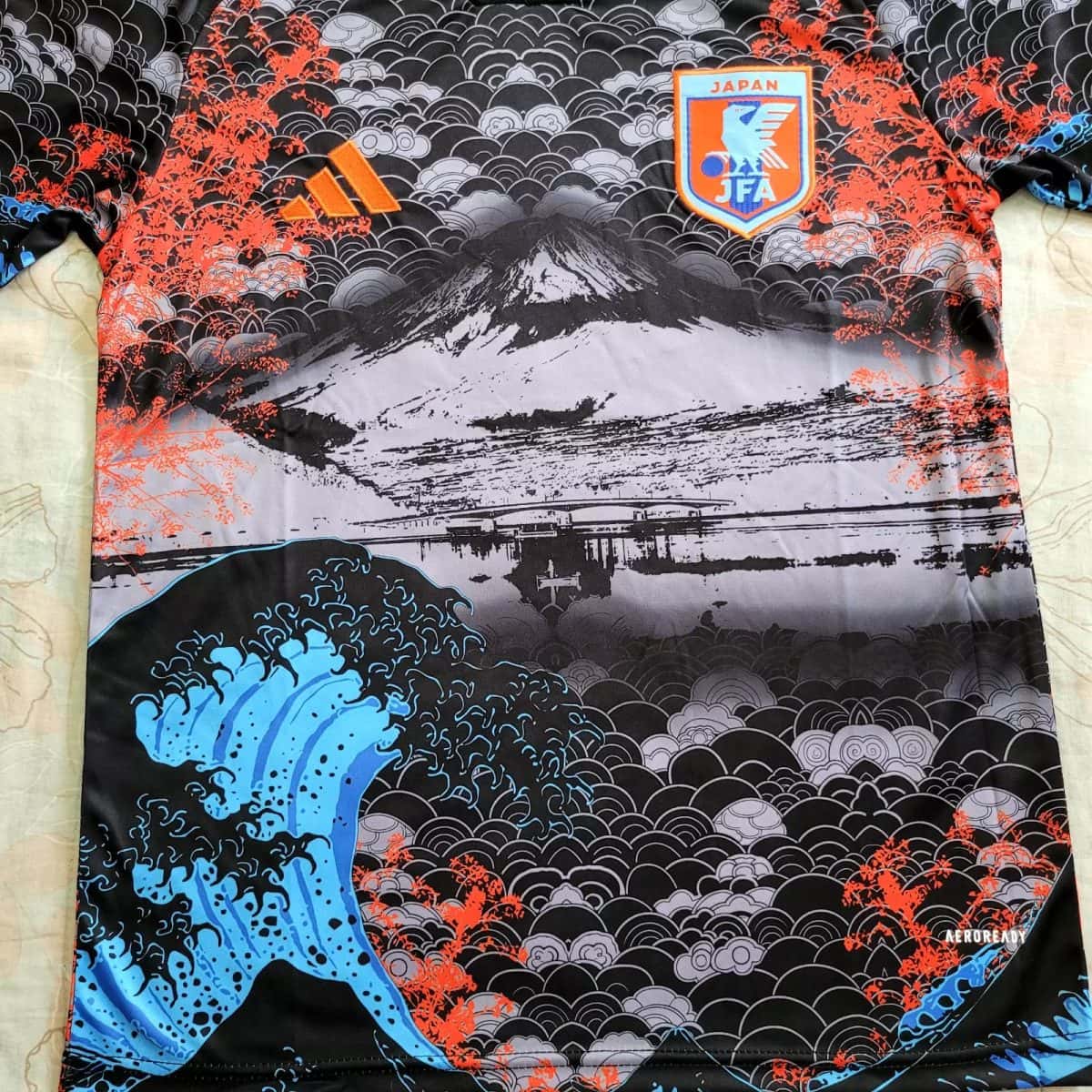 Sports jersey featuring black and gray cloud-wave pattern with orange accents, a blue wave motif, and a Japan Football Association crest.