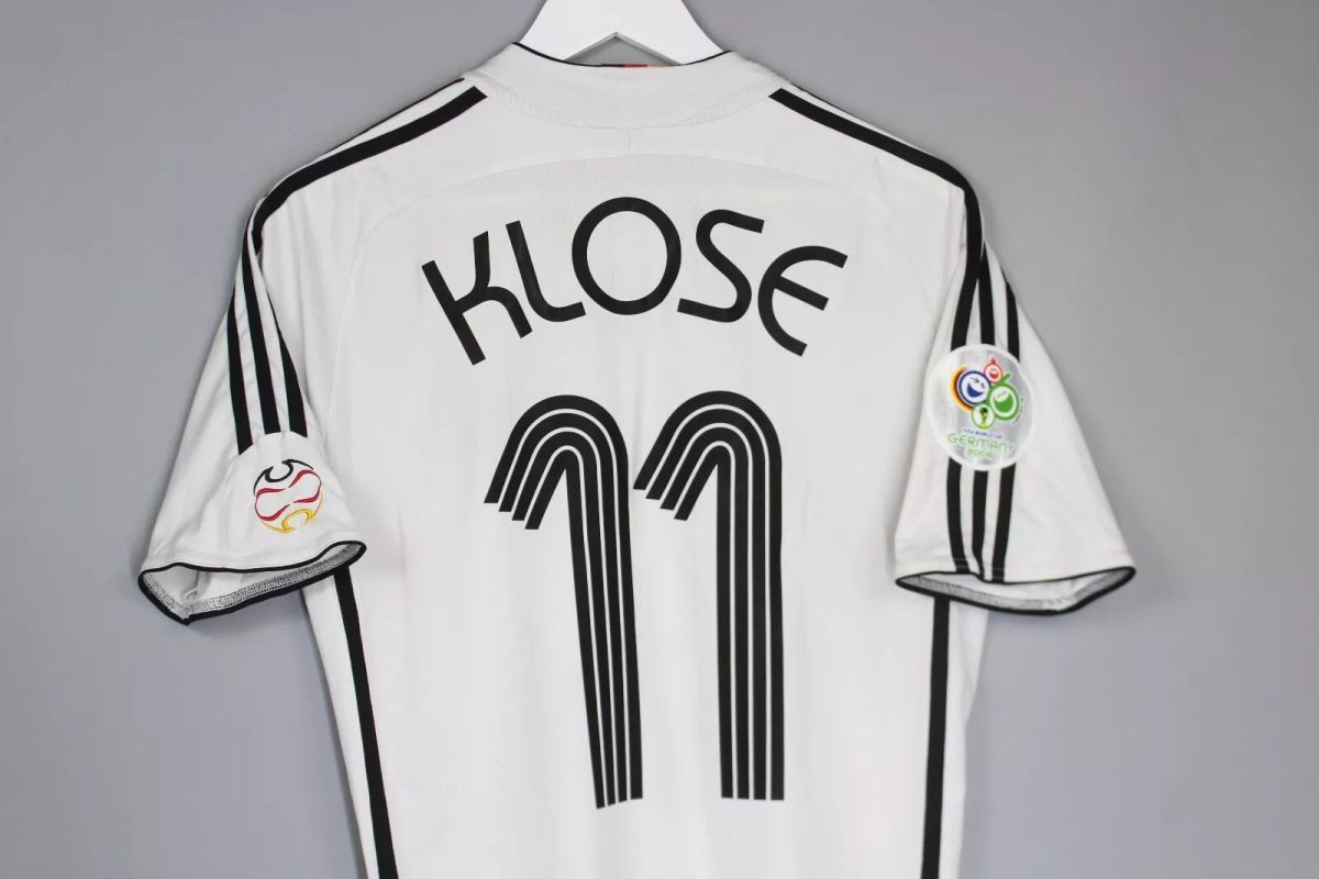 Back of white soccer jersey reading 'KLOSE' with a large black 11, hanging on a hanger; patches on the sleeves are visible.