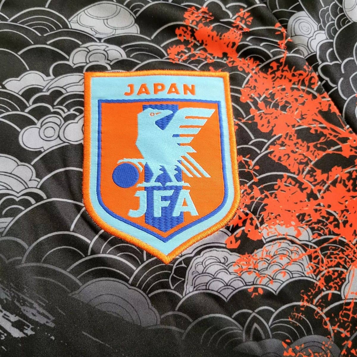 Patch showing Japan Football Association (JFA) emblem on light blue and orange, with 'JAPAN' at top, sewn onto patterned fabric background.