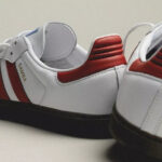 Pair of white Samba sneakers with red accents and a black sole on a light surface.