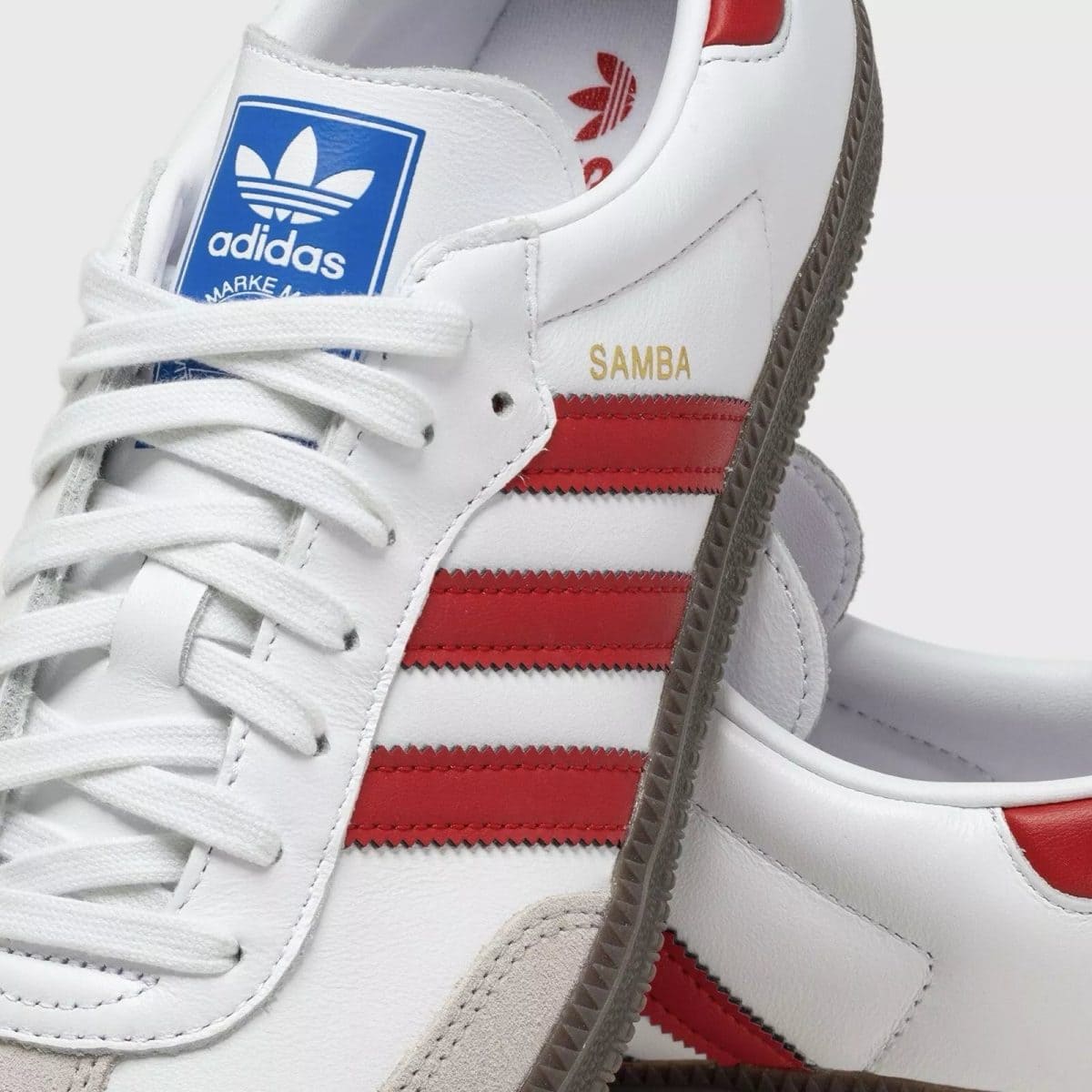 Close-up of a white Adidas Samba sneaker showing red stripes, blue tongue label, and white laces.