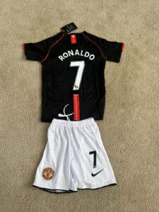 s-l1600_bc582931-b436-42fc-8b44-1423a2867c18 | UAE SPORT STORE Flat lay of a Manchester United Ronaldo #7 kit: black jersey with red piping and white shorts with the number 7 on a beige carpet.