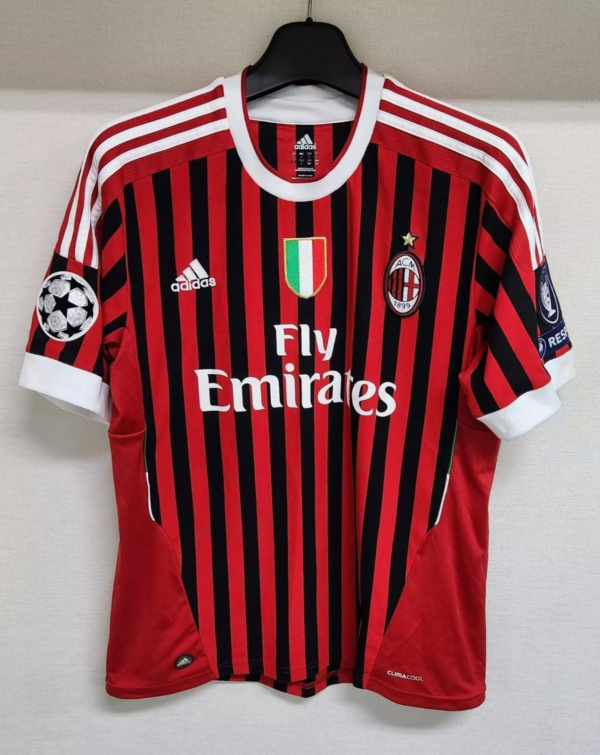 AC Milan home soccer jersey with red and black vertical stripes, Fly Emirates sponsor, club crest, and Italian flag shield on the chest, plus UEFA Champions League patch on the sleeve.