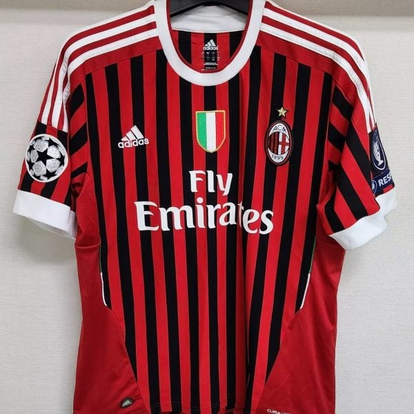 AC Milan home soccer jersey with red and black vertical stripes, Fly Emirates sponsor, club crest, and Italian flag shield on the chest, plus UEFA Champions League patch on the sleeve.