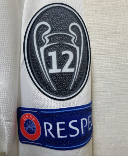 s-l500_15_08b562d8-6659-4480-9f5a-0b41170c2bd1 | UAE SPORT STORE Close-up of a white football jersey sleeve with a black shield badge displaying the number 12 and a blue band reading UEFA RESPECT.