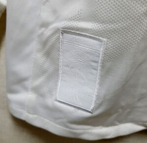 s-l500_17_1da9c053-240a-4658-a991-ae1dd8eddd45 | UAE SPORT STORE White fabric with a stitched rectangular label patch on the hem (blank embossed tag). Use of mesh-like texture visible nearby.