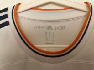 White Adidas Real Madrid jersey hanging on a hanger, close-up of the orange-trimmed collar viewable at the neck area.