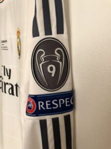 White soccer jersey sleeve with black stripes, displaying a circular trophy crest with the number 9 and a blue 'RESPECT' band; Real Madrid branding partially visible.