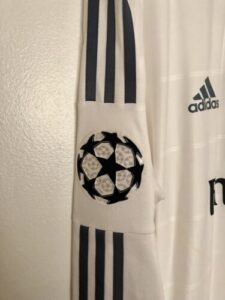 Close-up of a white Adidas soccer jersey sleeve with three navy stripes and a UEFA Champions League badge patch on the cuff.