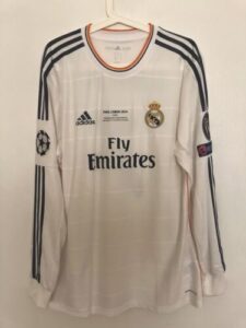 White Real Madrid football jersey with three navy stripes on shoulders and Fly Emirates logo on front, Real Madrid crest on chest, and Champions League patch on sleeve.